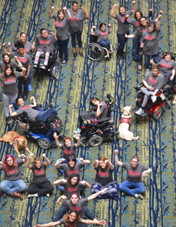 Thrive participants, some standing, some in wheelchairs, some with service dogs, shown from above, forming the women's symbol. Their arms up in the air, powerful.