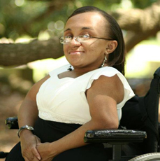 Black woman sitting in her wheelchair, smiling at the camera, wearing a black and white dress outside