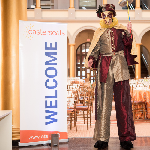 A person in costume on stilts next to a welcome sign.