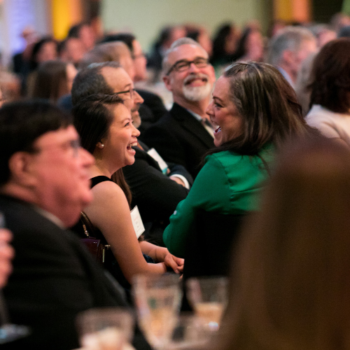 Two women seated next to each other laughing and smiling together amongst the rest of a seated crowd at the Advocacy Awards.