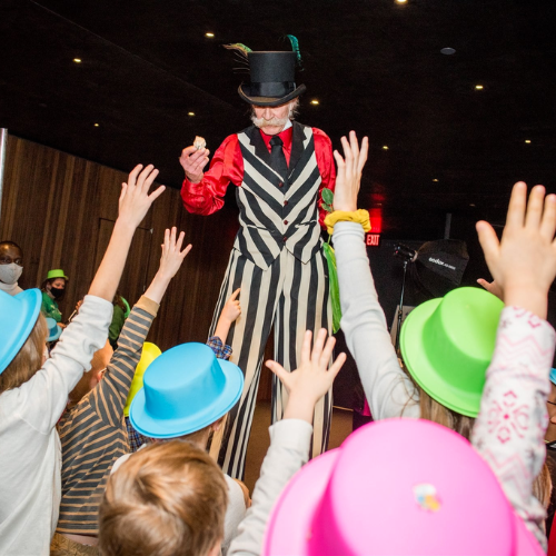 Man in a striped suit on stilts holding an object up in front of him as children gathered around him raise their hands.