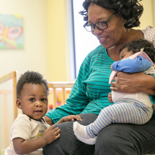 Teacher seated and holding one baby in her lap with another child standing at her knee in a classroom