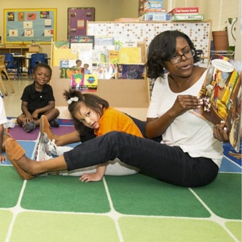A teacher reads a book to a group of students on a rug