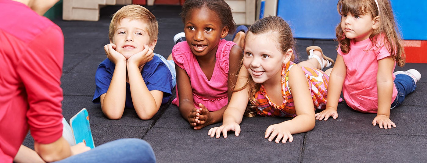 A teacher wearing a pink top is reading to four children. The kids are lying on the floor leaning on their elbows and smiling. 