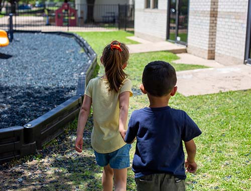 A little girl holding a little boy's hand as they walk toward a playground. They represent how children who attend Little Lonestar Academy can help each other learn and grow.