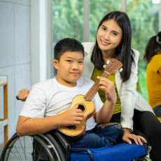 a boy who uses a wheelchair is holding a ukulele, a woman stands next to him and they smile at the camera