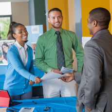 three people in business attire, a yellow wall in the background, one woman is handing papers to a man across from her