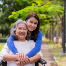 A woman using a wheelchair and her daughter hugging her from behind. Both women are smiling for the camera.