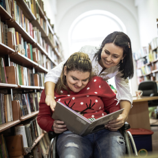 a woman using a wheelchair and another woman standing behind are reading a book together