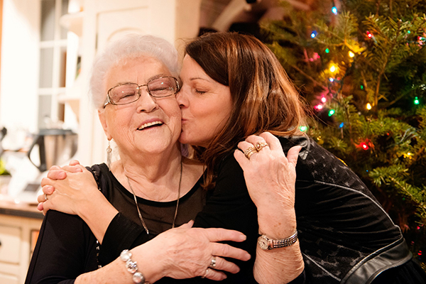 A middle age woman kissing an elderly woman in front of a Christmas Tree