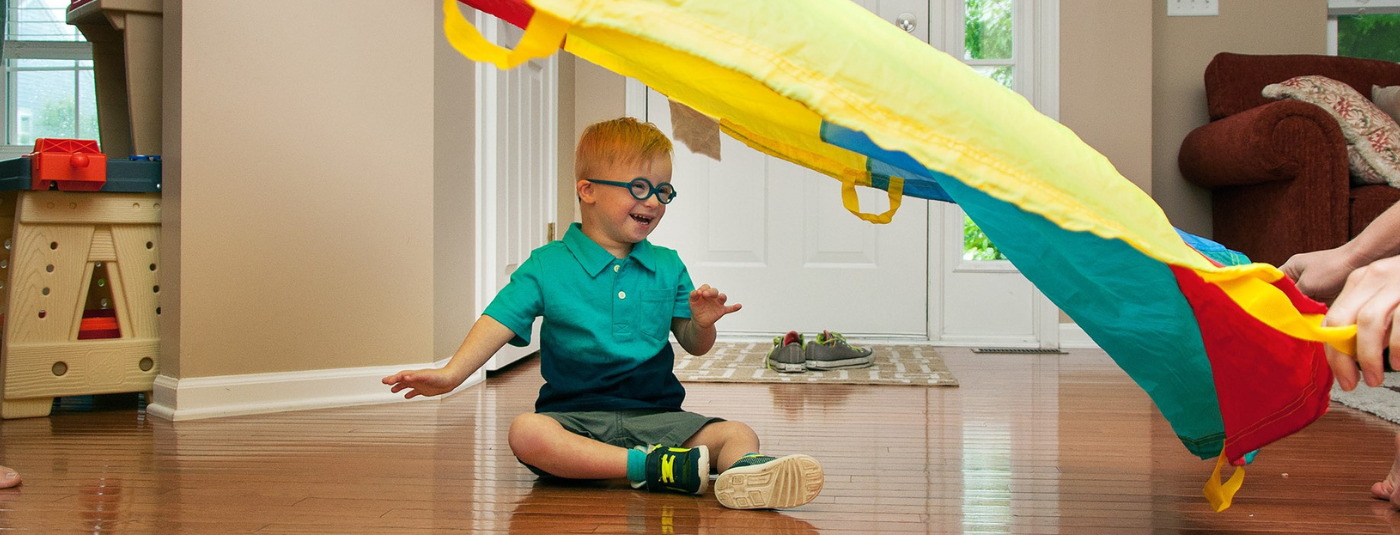 Toddler in blue glasses and ombre shirt smiling under a parachute