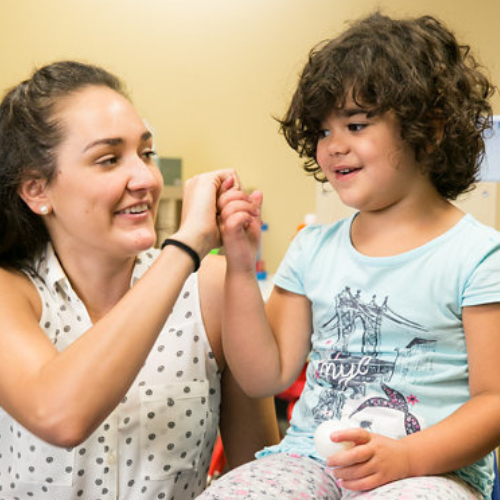 Smiling Woman Kneels and Holds Hands with Young Girl sitting in chair