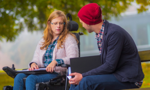 a young woman sits in an electric wheelchair on a college campus with a fellow student beside her