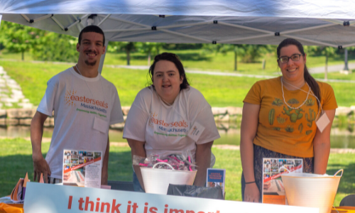 3 individuals stand under a tent at an outdoor event on a sunny day and smile.