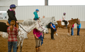 a group of people participate in adaptive riding lessons inside an indoor equestrian center