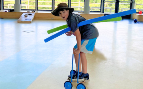 a boy rides a scooter inside a gym with noodles in his arms