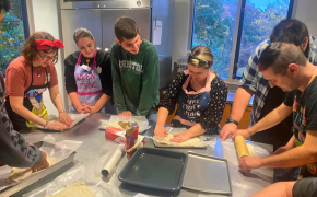 a group of young adults gather around a large kitchen prep table and prepare baked goods