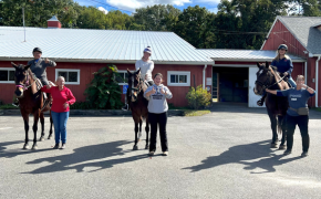 three horseback riders sit on their horses outside a barn, each with a leader standing next to them