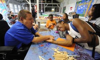 Two campers arm wrestle at a crafts table and smile