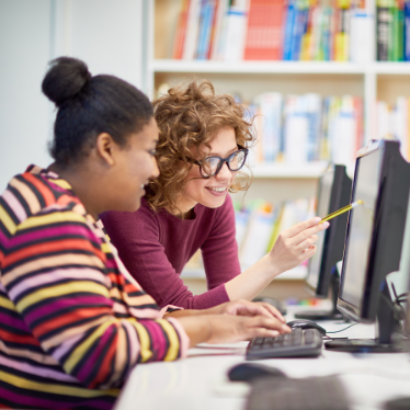 two women use a computer inside a library. One woman points to the screen with a pencil while the other watches