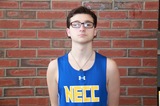 John Miller stands in his blue NECC track uniform in front of a brick wall