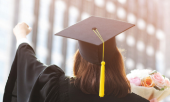 A graduate holds their fist up wearing a grad cap, and holding flowers with the other hand