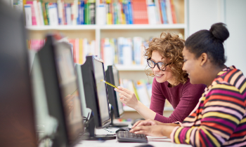 A woman helps another young woman on the computer in a library