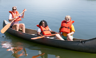 two campers with disabilities sit in a canoe paddled by a camp counselor on the water.