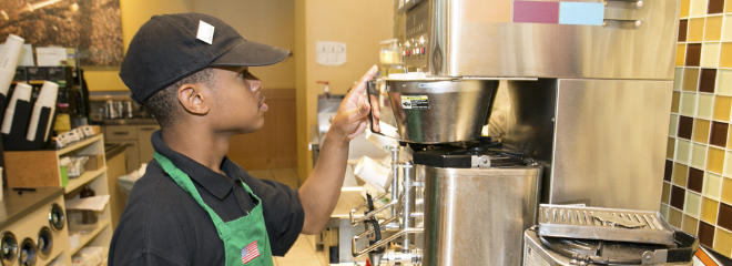 a young man with a disability is pictured working in a coffee shop, wearing a green apron while operating a coffee machine.