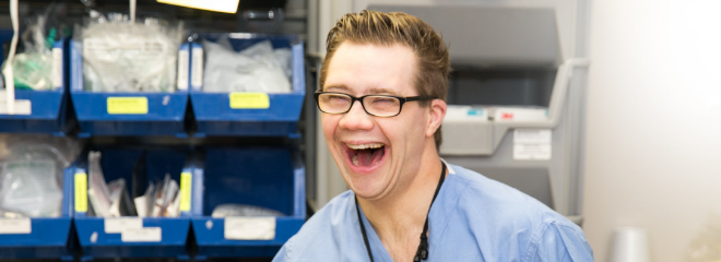 A man with a disability wears scrubs and smiles in a medical work setting.