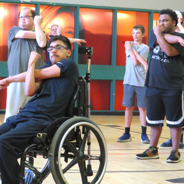 Young men participating in an accessible martial arts stretch inside of a gym.