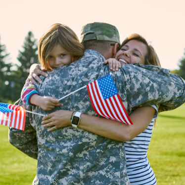 A solider wears his camo uniform and hugs his wife and daughter who are holding American flags