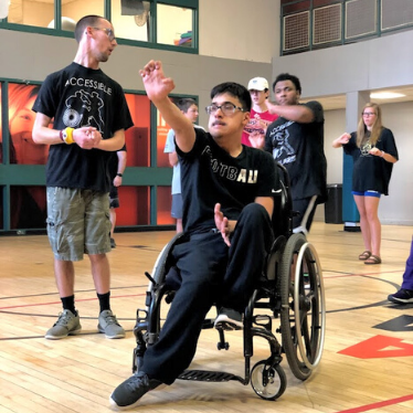 a young man sits in his wheelchair and participates in accessible martial arts exercise with his hand extended out