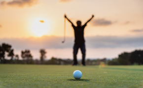 a golfer holds his arms up in the distance on a golf course with a golf ball in the front and center