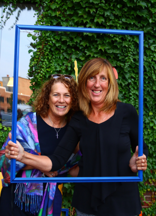 two women standing in front of an ivy wall holding up a blue frame and smiling together