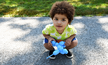 A child squats close to the ground outside holding a blue balloon animal