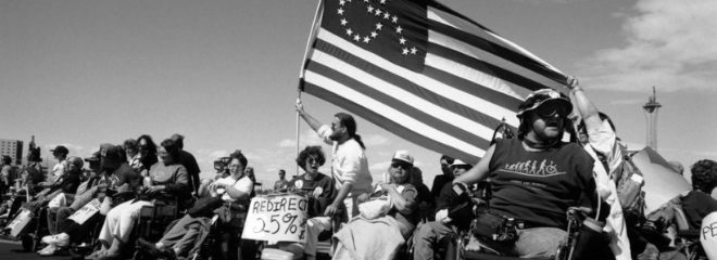 black and white image of people standing and sitting in wheelchairs, holding up a large American flag with a wheelchair symbol on it