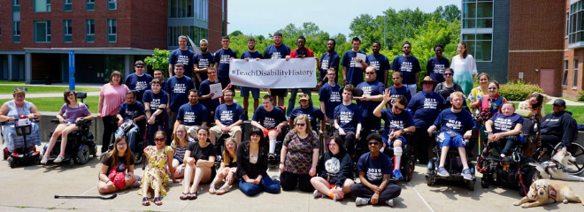 a large group of people sit/stand together and smile at the camera, holding a sign that reads #TeachDisabilityHistory