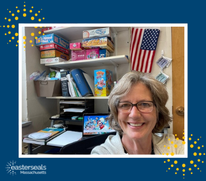 Doris, SLP, smiles in her office, with a stack of games/tools used during services behind her