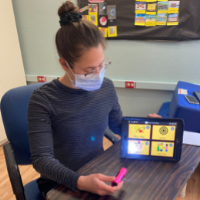 Cara sits in a classroom wearing a face mask, and uses an iPad to provide speech language pathology services
