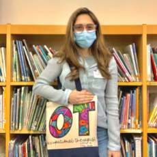 Liz stands with a face mask on inside a library setting, holding a tote bag that reads, 