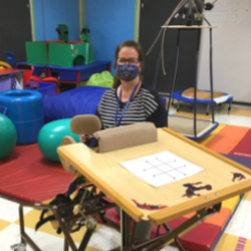 Paula sits inside a day habilitation center, behind a piece of rehabilitation equipment, with a face mask on