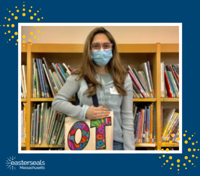 Liz stands with a face mask on inside a library setting, holding a tote bag that reads, "OT, Occupational Therapist"