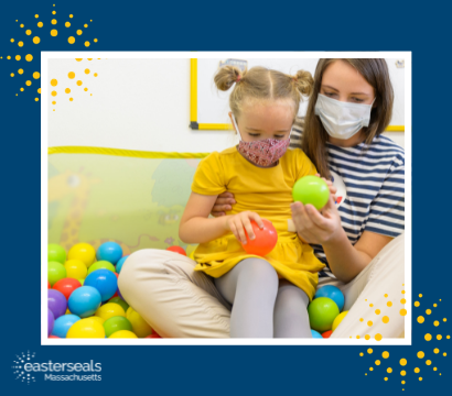 An occupational therapist sits with a young girl in her lap, both wearing face masks, while they both hold a ball in their hands