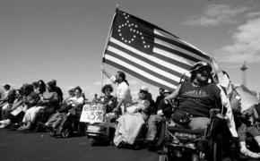people in wheelchairs protest together while flying an American flag with a wheelchair symbol