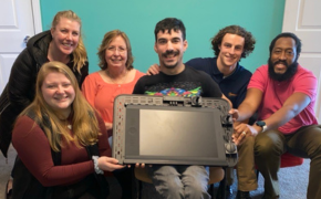 Alex and the AT team hold up a custom AT device that allows him to control his laptop with his foot
