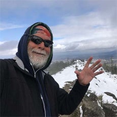 Connecting Communities participant, Gerardo B. He is on top of a snowy mountain, looking into the camera. He is smiling and waving his hand at the camera. He has sunglasses, a bandana, and a hooded sweatshirt on, and he has a gray beard.