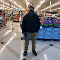 Dany is standing, inside of the Safeway grocery store, where he works. He is wearing a mask, a black jacket, and tan pants. His hands are in his pockets, and he is looking into the camera.