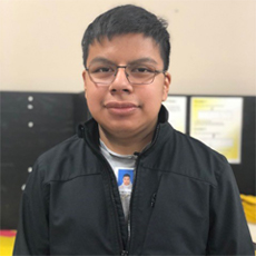Connecting Communities Participant, Eduardo Sanchez, looking directly into the camera for a headshot. He is a young, Latino male with black hair, glasses, and a black jacket on. He is in our Beaverton Connecting Communities office.