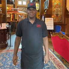 Connecting Communities Participant, Kevin. He is standing in the dining are at his work: The Old Spaghetti Factory. He's wearing his all-black uniform, which includes an apron and a hat. He's smiling and facing the camera.